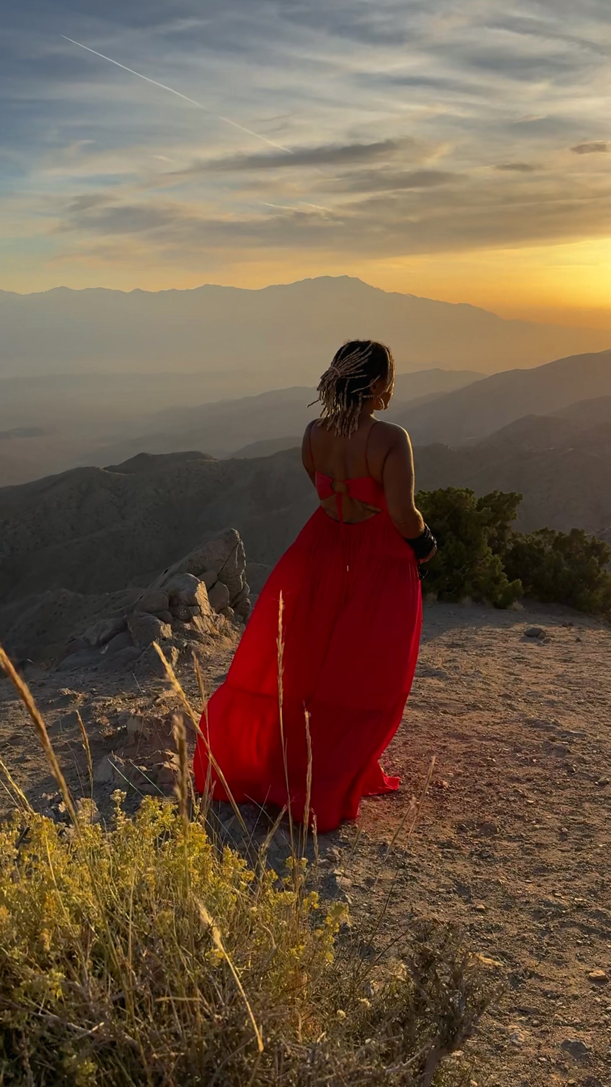 Jhaynane standing in a red dress above a mountain landscape at sunset during production.
